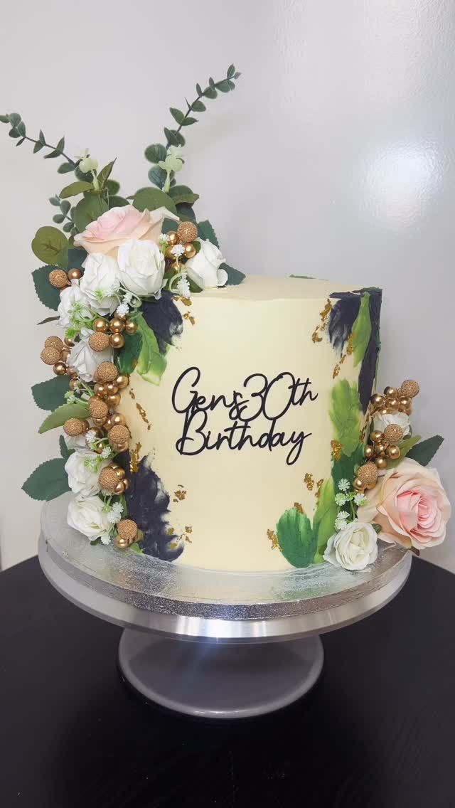 A white celebration cake with fresh flowers and gold lettering, made by Becca